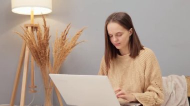 İç işleri için not defteri. Evde dizüstü bilgisayarla çalışıyor. Düşünceli düşünceli kadın bej kazak giyiyor dizüstü bilgisayar kullanıyor oturma odasında otururken uzaktan projeyi düşünüyor.