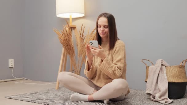 漂亮的白种人女人 穿着米黄色毛衣 坐在家里的地板上浏览互联网网页 开心地笑着 — 图库视频影像