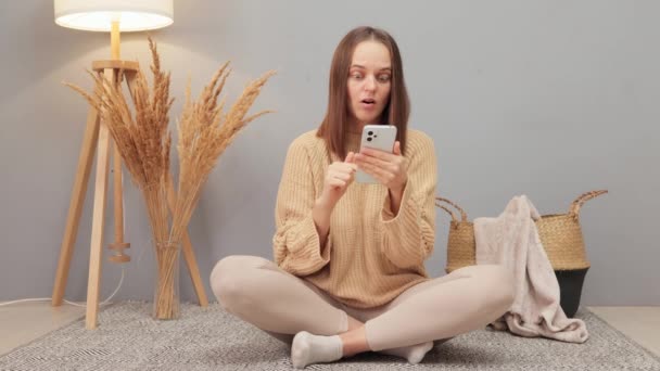 スマートフォンで女性チャット 驚くほど衝撃を受けた大人の白人女性は 携帯電話のブラウジングを使用してベージュセーターを着用し 家庭内インテリアの床に座っている間に驚いた顔でページを表示します — ストック動画