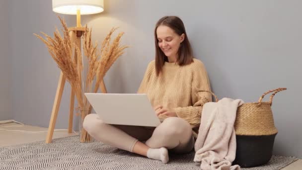 オンラインでの就職機会 リモートワーク接続のヒント 起業家技術について リビングルームの床に座っている間 ラップトップを使用してベージュセーターを身に着けている喜ばしい笑顔の女性 — ストック動画