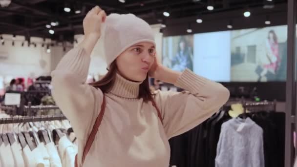 ファッションショッピングを楽しむ寒い季節のための冬の帽子を選ぶファッションストアの鏡の前で冬の服をしようとする若い美しい女性 — ストック動画