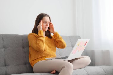 Sanal bir çalışma alanında serbest çalışan. Evdeki sosyal medya ağı. Kafkasyalı bir kadın dizüstü bilgisayar kullanıyor. Evdeki koltukta oturuyor. Baş ağrısı çekiyor.