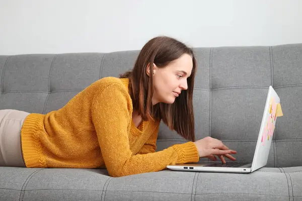 Internet browsing for work or leisure. Laptop work for freelancers. Brown haired Caucasian woman wearing orange sweater using laptop while lying on sofa at home
