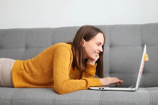 Tech freelancer job remote. Social media network. Leisure entertainment and watching. Cute charming adorable Brown haired woman wearing orange sweater using laptop while lying on sofa at home