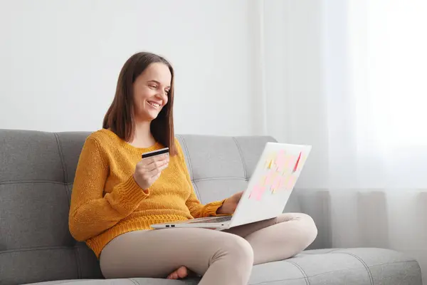 Internet banking transactions. Credit card purchases. Smiling happy delighted Caucasian brown haired woman working with laptop and credit card at home while sitting on sofa