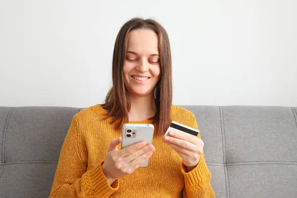 Phone credit pay. Transaction cellphone ecommerce. Cheerful joyful woman wearing orange jumper using mobile phone and holding credit card while sitting on sofa at home