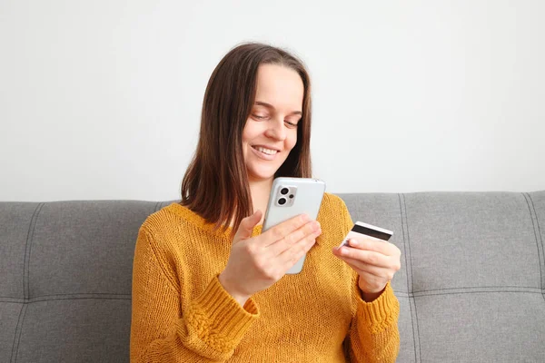 Online shopping experience. Mobile payment method. Attractive positive woman wearing orange jumper using mobile phone and holding credit card while sitting on sofa at home