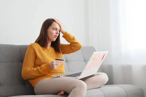 Technology and financial transactions. Shocked Caucasian brown haired woman working with laptop and credit card at home while sitting on sofa having troubles with online transaction