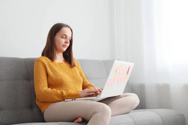 Home-based business with technology. Remote work and laptop usage. Online education and e-learning. Sad Caucasian woman using laptop sitting on sofa at home