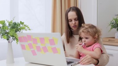 Serbest ve annelik. Evde dizüstü bilgisayarla çalışan ve bebeğini kucağına alan ve mutfakta bilgisayar kullanan genç bir kadın.