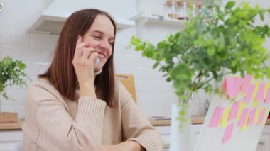 Çekici, neşeli, beyaz saçlı, uzaktan kumandalı bilgisayar kullanan, evinde oturmuş, cep telefonuyla konuşan, neşeli, kahverengi saçlı bir kadın.