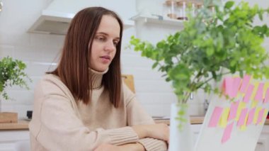 Uzaktan kumanda için dizüstü bilgisayar kullanan başarılı bir Kafkasyalı kadın. İnternette konuşan, mutfakta oturan, internette konuşan bir kadın.