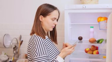 Kafkasyalı bir kadın markete gitmek için akıllı telefonunu kullanırken buzdolabında yiyecek siparişi verirken buzdolabından yemek hazırlamak için ürünler alıyor.