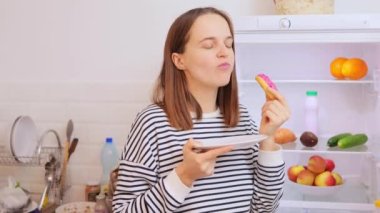 Tatmin olmuş bir kadın buzdolabının yanında sağlıksız yiyecekler yiyor. Elinde donutla tatlı atıştırıyor. Sağlıksız beslenmenin tadını çıkarıyor.
