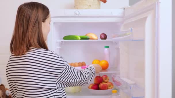 キッチンオープニング冷蔵庫でストライプシャツを着ている空腹の白人女性は 甘いドーナツを食べることを取ります 不健康な栄養肥満ダイエットの失敗過剰な食品中毒 — ストック動画