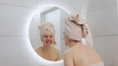 Kafasına havlu sarılmış neşeli beyaz kadın banyoda durup yüzüne dokunan aynaya bakıyor görünüşüne bakıyor.