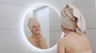 İyi olmak rutin bir şey. Bayan cilt bakımı. Tazelik bakımı. Kafkasyalı kadın kafasına havlu sarılmış. Yüz temizleme fırçası kullanıyor. Banyoda duran aynaya bakıyor.