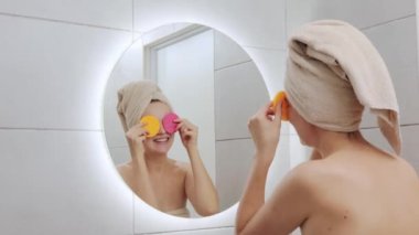 Kafasına havlu sarıp gözlerini süngerle kaplayan komik beyaz kadın sabahları cilt bakımını yaparken banyodaki yansımasına bakarak eğleniyor.