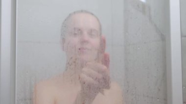 Attractive young woman drawing heart on weeping glass shower door enjoying rest in douche washing her body standing behind steam blurred glass with water drops