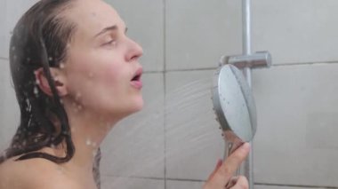 Genç, beyaz, kahverengi saçlı, çıplak kadın duş başlığı kullanarak kabinde şarkı söylüyor. Banyo güzellik tedavisinde günlük vücut bakımı işlemlerinin keyfini çıkarıyor.