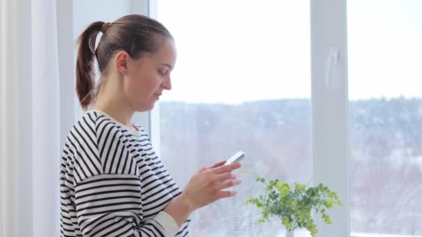 Odaklanmış Kafkasyalı Kadın Cep Telefonu Ile Evinin Penceresinin Yanında Duruyor — Stok video