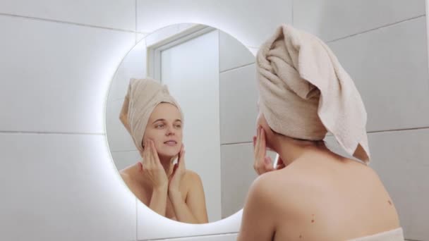 笑顔のコーカサス人女性は 浴室に立っている化粧品治療のためにクリームを使用した後 彼女の反射で彼女の顔をマッサージする鏡を見ながら彼女の頭にタオルで包まれました — ストック動画