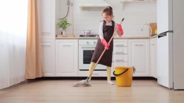 Ev eldivenli küçük bir kız mutfak yakınında modern paspas ve su kabıyla yerleri yıkıyor ev temizliği yapıyor mikrofon olarak fırçayı kullanıyor annesine ev işlerinde yardım ederken eğleniyor.
