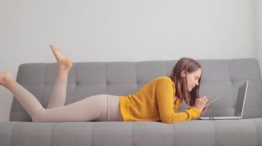 Evde oturma odasındaki kanepede uzanmış bilgisayar başında cep telefonuyla internet sayfalarında gezinen bilgisayarları yazarken bilgisayarını kullanan Kafkasyalı serbest çalışan bir kadın.