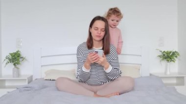 Sevgi ve mutluluğu ifade eden anne ailesinin yanında zıplayıp duran küçük kızıyla vakit geçirirken akıllı telefon kullanan sevimli bir kadın.