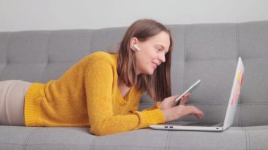 Oturma odasındaki kanepede uzanmış dizüstü bilgisayar kullanan çekici, genç, beyaz kadın elinde akıllı telefonla internet işini birleştiriyor ve cep telefonuyla sosyal ağları kontrol ediyor.