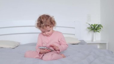 Küçük bebek dalgalı saçlı sarışın kız yatakta oturuyor ve akıllı telefon kullanıyor izlemek için çizgi film seçiyor ya da oyun oynamak için çocuk oyuncağı.