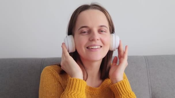 迷人的黑发女性穿着黄色毛衣 头戴耳机 一边欣赏音乐 一边在家里的沙发上休息 一边欣赏最喜欢的Mp3曲目 — 图库视频影像