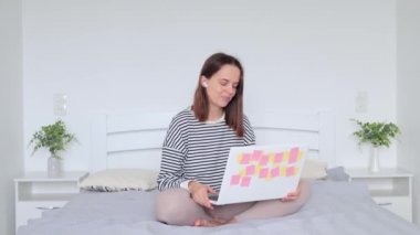 Yatakta dinlenen bir kadın bilgisayar kullanarak samimi yatak odasında oturmuş kulaklıklarıyla el sallıyor. Arkadaşına çevrimiçi sohbetin keyfini çıkarırken el sallıyor.
