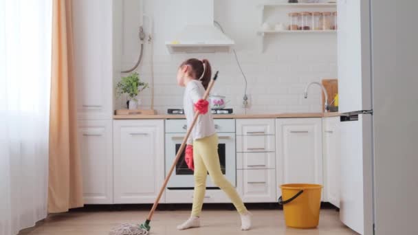 可悲的小女孩 戴着马尾辫 戴着保护乳胶手套 用现代拖把在厨房附近用可移动的刷子擦拭地板 做她的家务活 — 图库视频影像