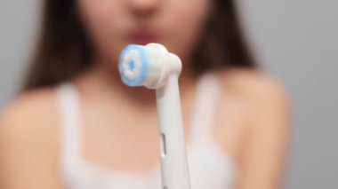 Tanımlanamayan kahverengi saçlı küçük bir kız dişlerini elektrikli diş fırçasıyla fırçalıyor. Gri arka planda izole edilmiş. Günlük diş bakımı için ağız bakımı ekipmanları tutuyor.