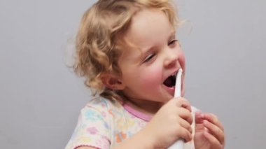Dalgalı saçlı, dişlerini elektrikli diş fırçasıyla fırçalayan, gri arka planda izole edilmiş, günlük diş bakımı için oral bakım malzemesi taşıyan bebek.