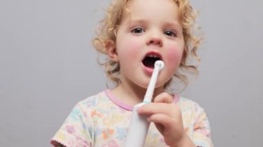 Bebek diş koruması. Ufaklığın sağlığı. Kadın fırçalama rutini. Sarı dalgalı saçlı, dişlerini elektrikli diş fırçasıyla fırçalayan, tatlı bebek. Gri arka planda izole edilmiş.