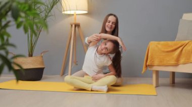 Sağlık için aile egzersizi. Kadın antrenör jimnastiğe rehberlik eder. Canlılık için ev zindeliği. Beyaz kadın ve küçük kızı evde spor yapıyorlar.