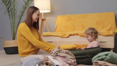 Aile tatili macerası. Anne ve bebek seyahati. Neşeli toplanma anları. Küçük kızıyla seyahat etmek için bavulunu hazırlayan mutlu beyaz kadın evdeki oturma odasında