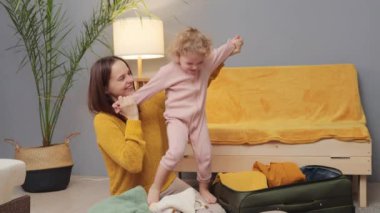 Aile tatili planlaması. Ufaklıklarla seyahat etmek. Çocuklar için neşe paketliyorum. Beyaz kadın, küçük kızıyla birlikte seyahat için bavulunu hazırlıyor. Oturma odasında.