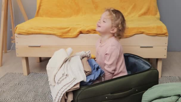 陽気なブロンドの茶色の毛髪の小さな赤ん坊の少女はスーツケースに座って楽しい笑顔で楽しい旅の準備をしている間 母親は喜んで服を荷造りしています — ストック動画