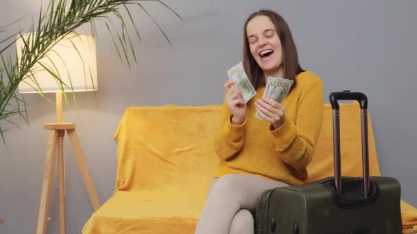 非常に幸せなポジティブな女性は ドル紙幣とソファに座ってカジュアルな服を着て 旅行のために彼女の貯金を費やして彼女の旅行ダンスを楽しんでいます — ストック動画