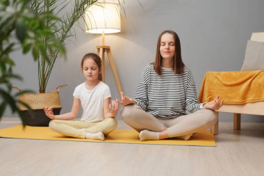 Aile yogası. Güzel genç bir kadın ve sevimli küçük kızı evde yoga yapıyorlar. Lotus pozisyonunda oturma odasında oturuyorlar. Anne çocuklara meditasyon yapmayı öğretiyor.