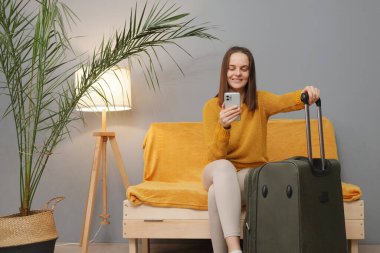 Gülümseyen Kafkasyalı, kahverengi saçlı, turuncu kazaklı, koltukta oturan, koltukta oturan, taksiyi bekleyen, SMS yollayan, seyahat etmeye hazır bir kadın.