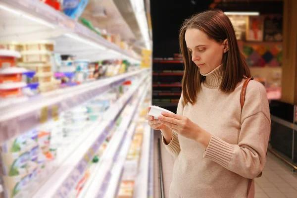 Young Caucasian brown haired woman buying diary product and reading food label in grocery store wearing casual sweater reading information about product expiration date