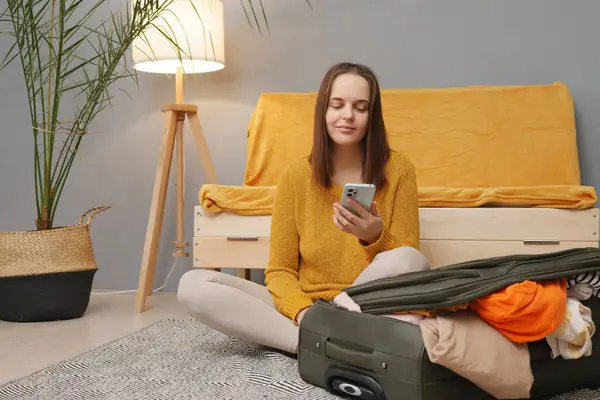 Stylish tourist lifestyle. Packed for a journey. Caucasian young adult woman packing suitcase at home using mobile phone booking clothing for traveling