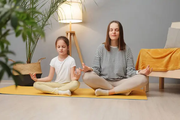 Family yoga. Beautiful young woman and her charming little daughter doing yoga together at home sitting in lotus position on floor in living room mom teaching child to meditate
