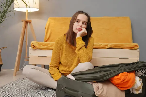 Bored Caucasian young adult woman packing suitcase at home taking too much attire waiting for her departure with sad boring facial expression leaning on her face looking at camera