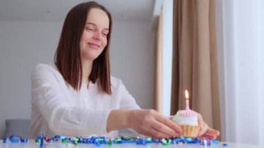 Evde doğum günü eğlencesi. Kafkasyalı genç bir kadın doğum günü pastası ile masada oturmuş yanan mum üflüyor ve tek başına aydınlık odada kutlama yaparken dilek diliyor.