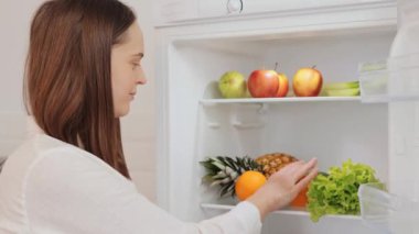 Kahverengi saçlı kadın buzdolabı rafları düzenliyor buzdolabından taze portakallar alıyor lezzetli kokan aroma lezzetli sağlıklı eroin için meyve seçiyor.